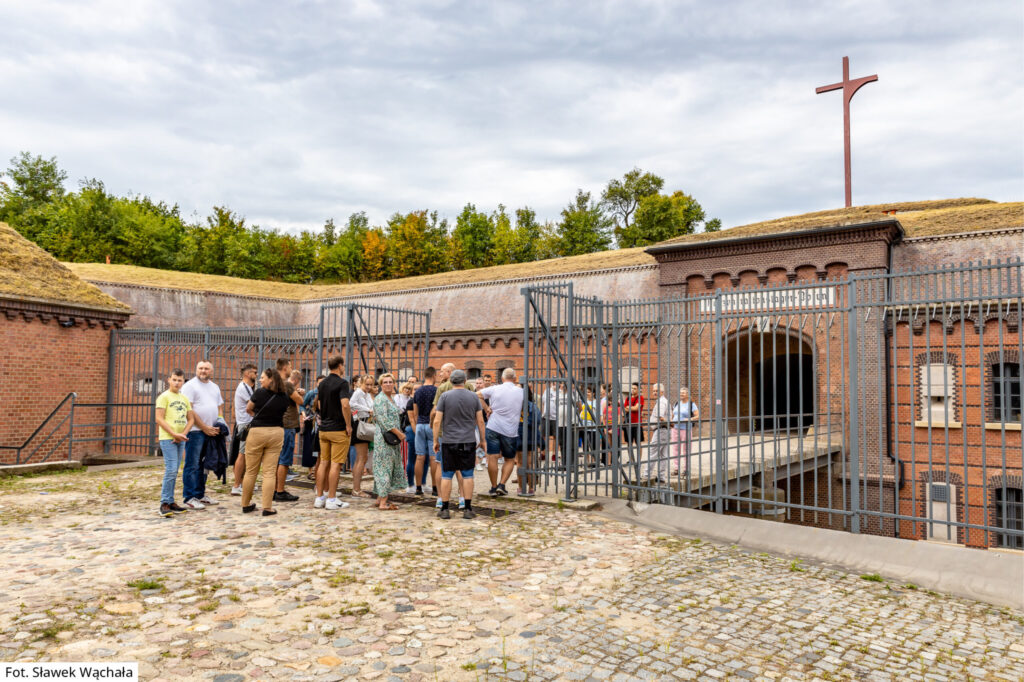 Grupa zwiedzających stoi na moście przed wejściem do Fortu VII. W tle po prawej krzyż.